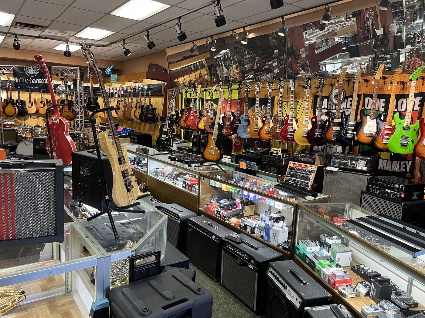 Interior of B & B Loan Company in Fort Wayne, showing a wall of dozens of used electric and acoustic guitars hanging above a row of glass jewelry and effects-pedal display cases, with KISS and Bob Marley posters on the back wall.
