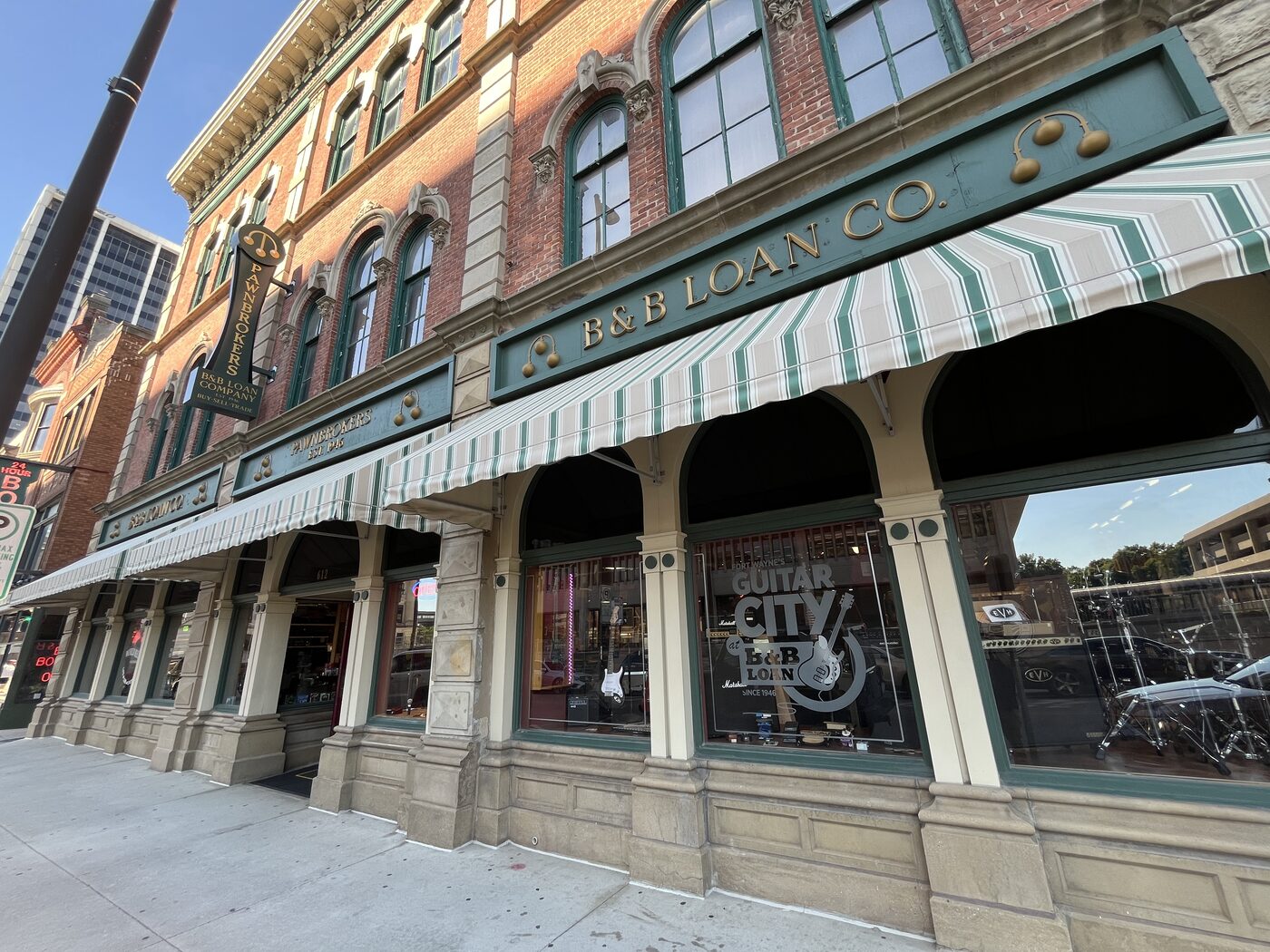 Historic two-story brick storefront of B & B Loan Company in downtown Fort Wayne, Indiana, with a green-and-cream striped awning, gold &lsquo;B & B Loan Co.&rsquo; lettering on the fascia, a vertical &lsquo;Pawnbrokers&rsquo; blade sign, and &lsquo;Guitar City at B & B Loan since 1946&rsquo; window etching.